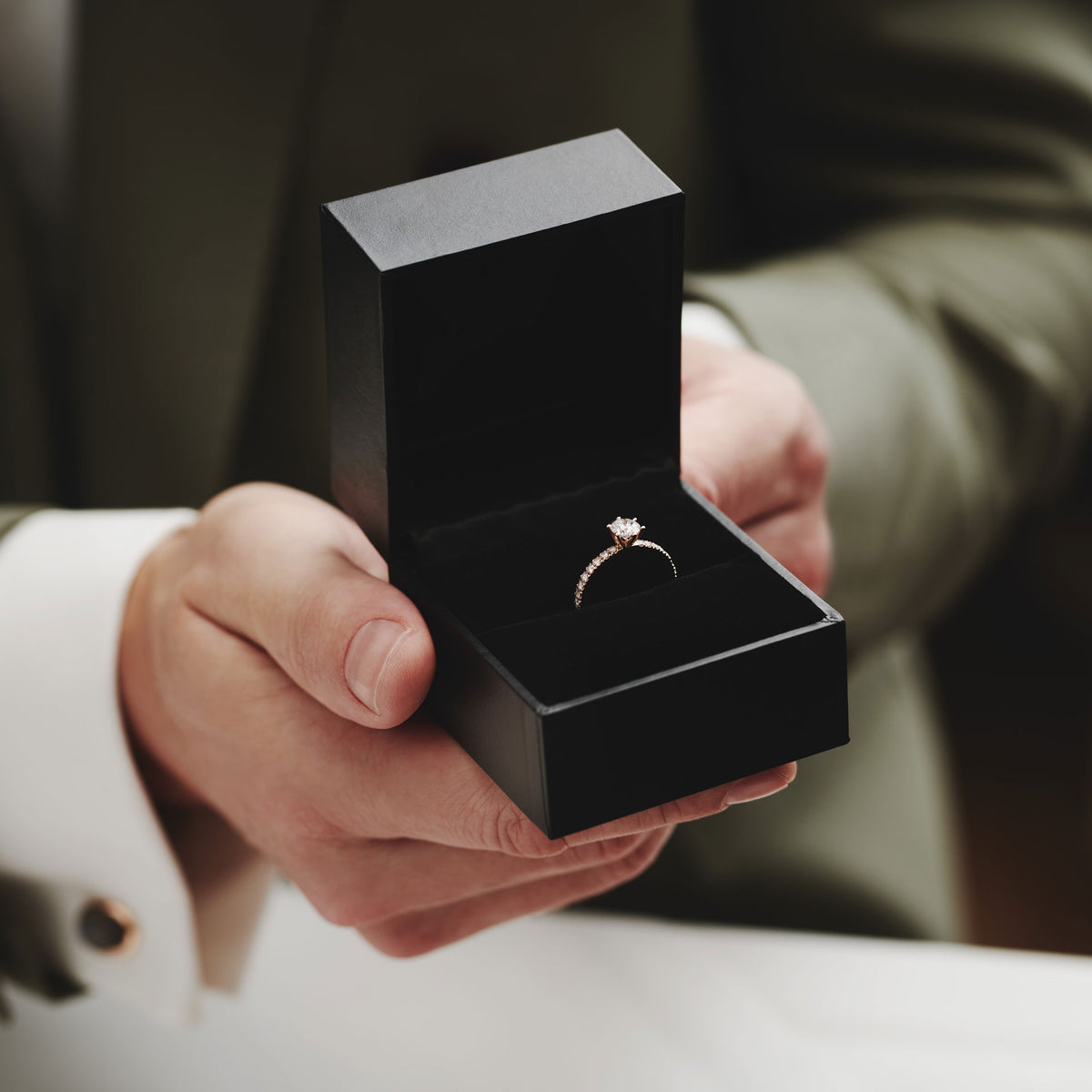 Eine Person im Anzug hält eine geöffnete schwarze Ringschachtel mit dem diamantenen Verlobungsring Schullin RING GRACE 0,25 CARAT in der Hand, der einen Heiratsantrag andeutet.