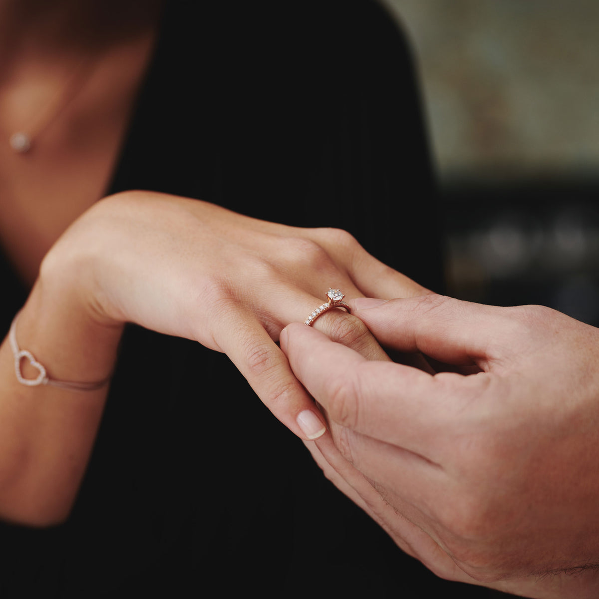 Eine Nahaufnahme von jemandem, der den Schullin RING GRACE 0,25 CARAT an den Finger einer anderen Person steckt, die ebenfalls ein Herzarmband trägt. Beide Hände berühren sich sanft.