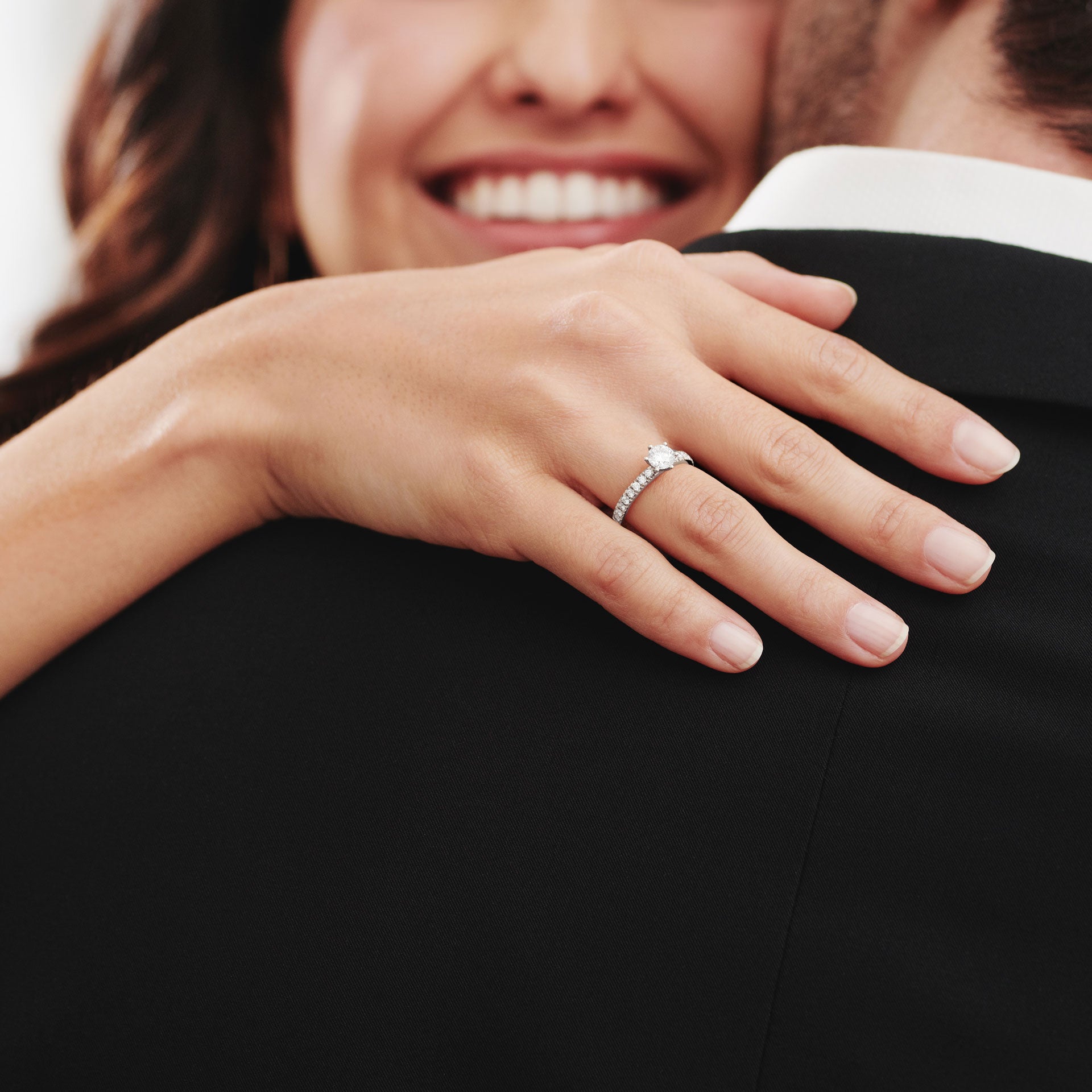Eine Frau lächelt, während sie einen Anzugträger umarmt und dabei den Schullin RING GRACE 0,75 CARAT Diamant-Verlobungsring an ihrer linken Hand präsentiert.