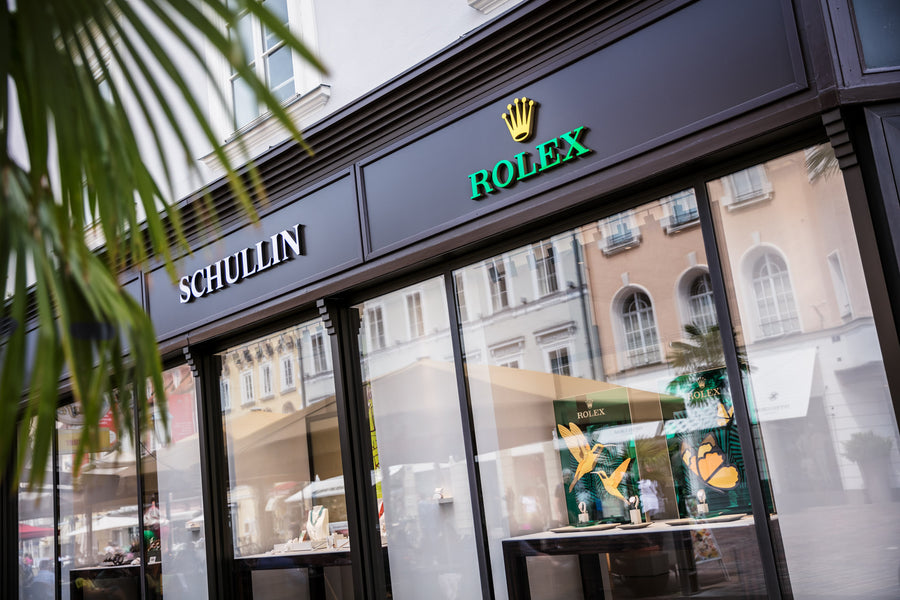 Eine Straßenansicht eines Schullin-Geschäfts in einem Stadtgebiet mit einer Rolex-Abteilung, in deren Schaufenstern Uhren ausgestellt sind. Das Gebäude hat eine dunkle Außenfassade mit großen Glasfenstern, in denen sich die umgebende Architektur spiegelt. Palmwedel rahmen die Szene teilweise ein.