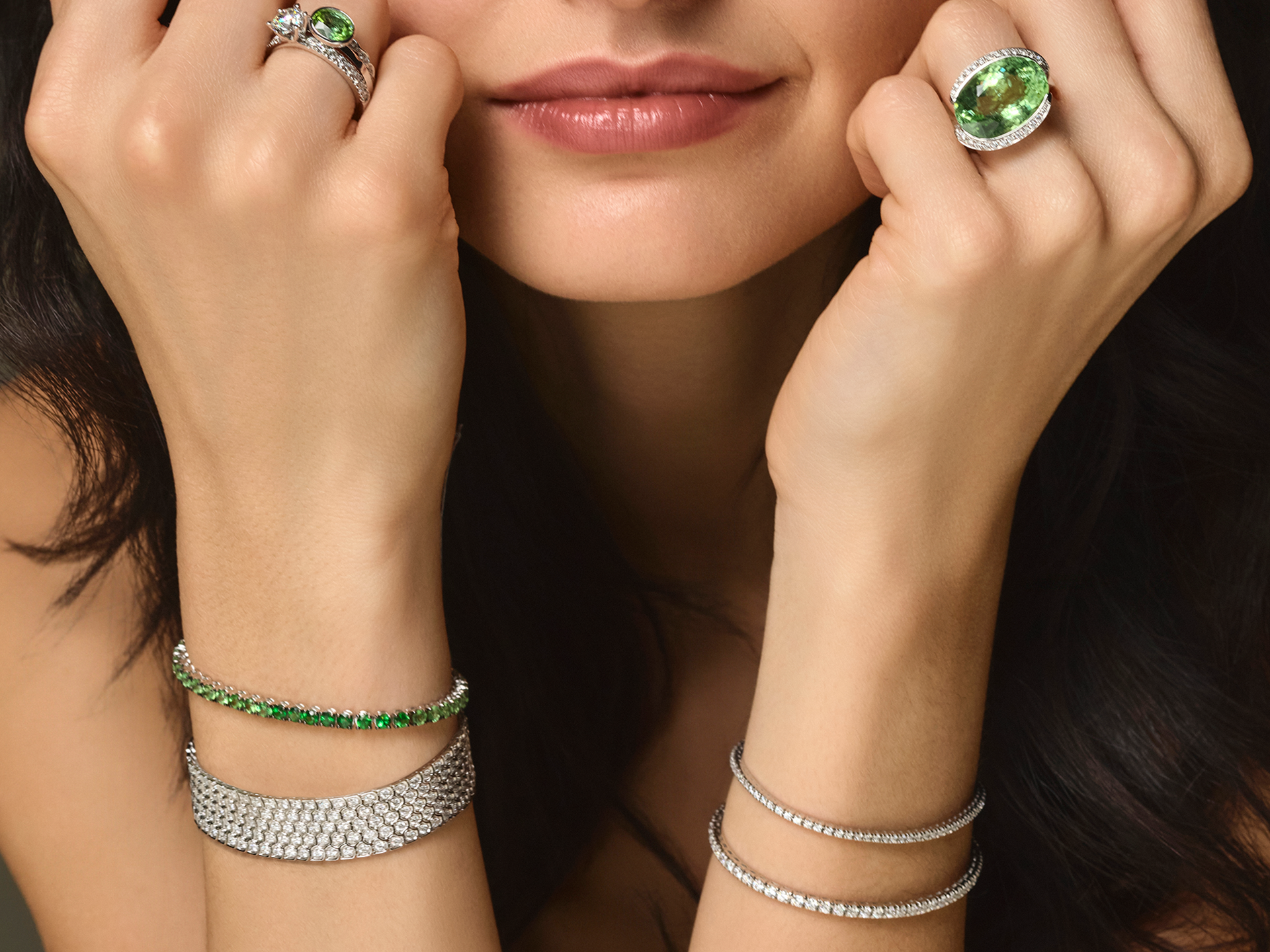Eine dunkelhaarige Frau trägt ein Schullin TENNISARMBAND 2,50 CARAT und auffällige grüne Edelsteinringe an beiden Händen in der Nähe ihres Gesichts, mit einem leichten Lächeln auf den Lippen.