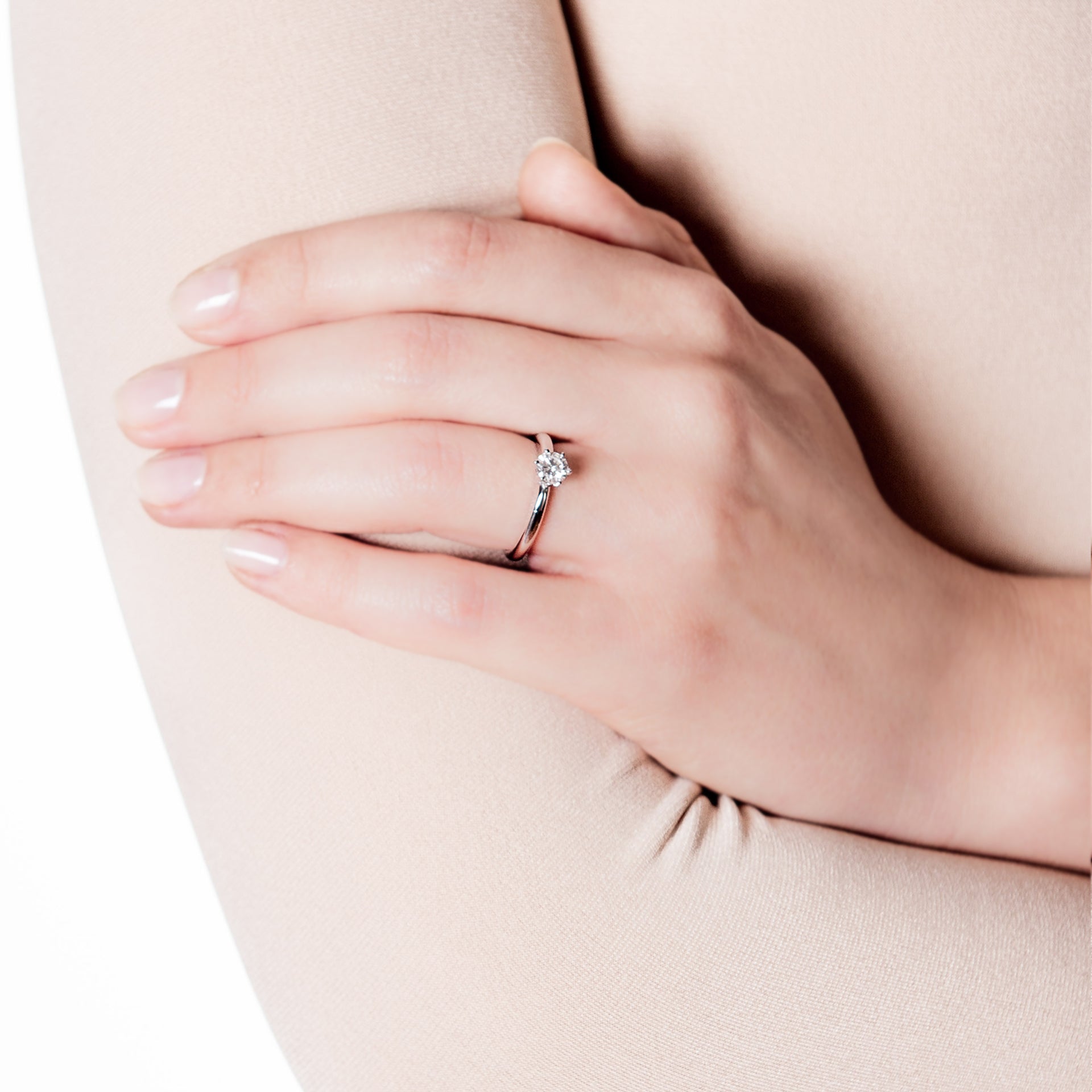 Eine Frauenhand mit hellrosa Nagellack ruht auf ihrem Arm und präsentiert den Schullin SOLITAIRE RING PRINCESS 0,45 CARAT - einen silbernen Verlobungsring mit einem einzelnen Diamanten im Prinzessinnenschliff, der vor einem weichen, neutralen Hintergrund gefasst ist.