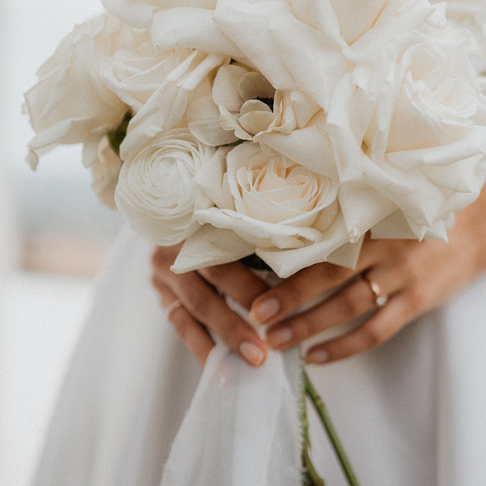 Eine Nahaufnahme einer Person, die einen Strauß weißer Rosen und Ranunkeln hält, der in weißen Stoff eingewickelt ist. Die mit einem Ring geschmückte Hand der Person umfasst sanft die Stiele. Der Hintergrund ist leicht verschwommen.