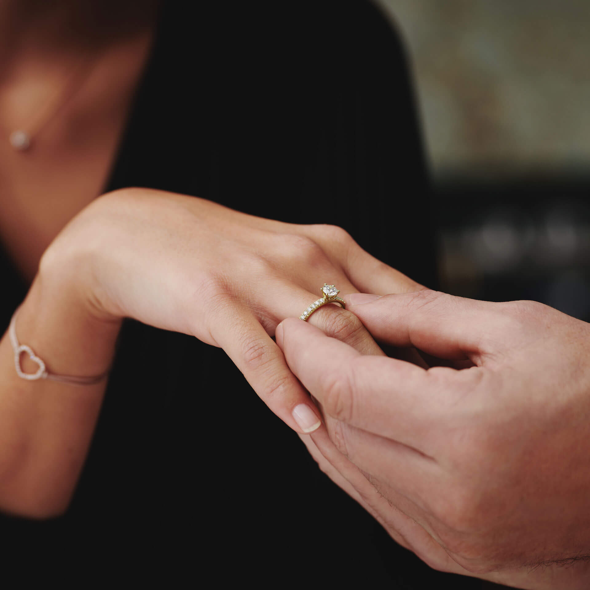 Eine Person steckt einer anderen den exquisiten Schullin RING GRACE 0,46 CARAT an den Finger. Der Gelbgoldring bringt den Solitär-Diamanten elegant zur Geltung, während die andere Person ein Armband mit einem Herzanhänger trägt. Der sanft verschwommene Hintergrund unterstreicht diesen intimen Moment auf wunderschöne Weise.