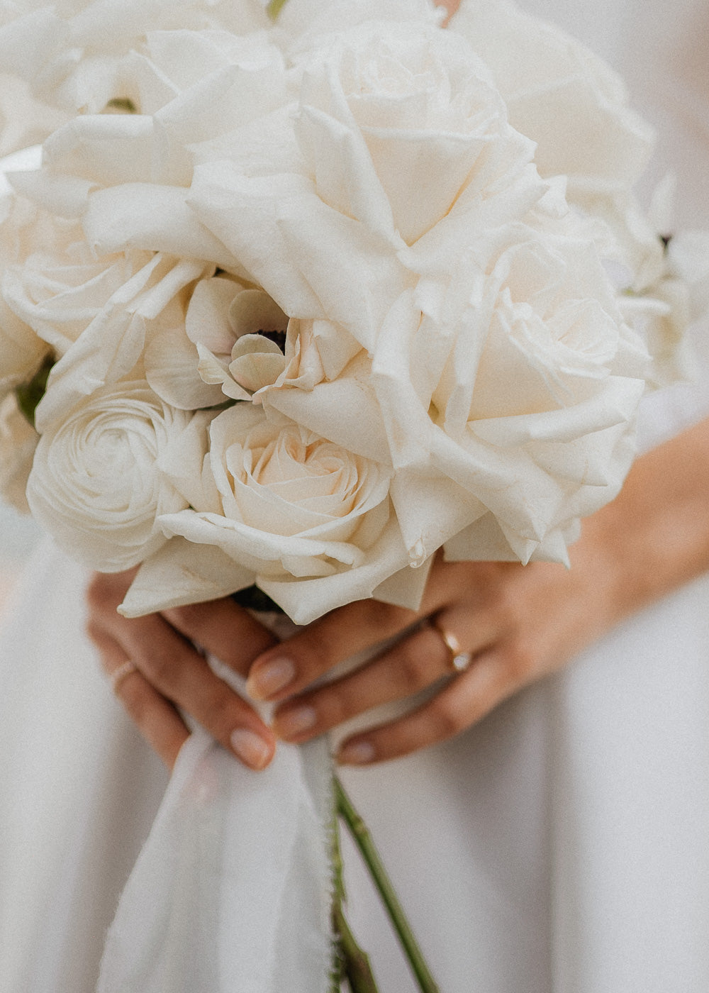 Eine Nahaufnahme einer Braut, die einen Strauß weißer Rosen und anderer Blumen hält. Ihre Hände sind sichtbar, eine davon trägt einen einfachen goldenen Ring. Der Strauß bildet einen Kontrast zu ihrem weißen Kleid und schafft eine elegante und ruhige Komposition.