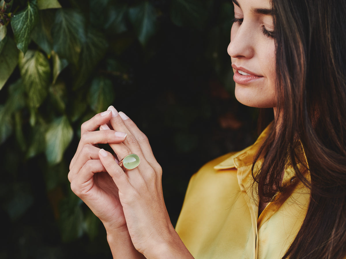 Eine Frau in einem gelben Hemd berührt sanft eine Hand mit einem großen grünen Ring an ihrem Finger. Sie steht vor üppigen grünen Blättern und betrachtet den Ring mit einem sanften Gesichtsausdruck.