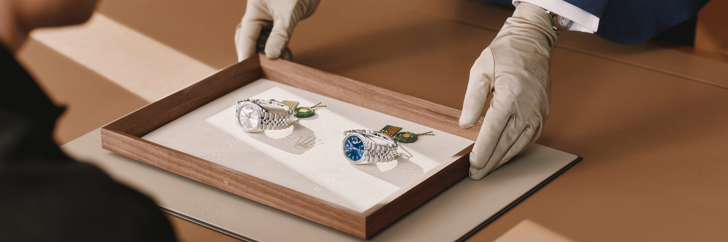 Eine Person mit Handschuhen überreicht einer anderen sitzenden Person auf einer braunen Fläche ein Holztablett mit zwei Luxusarmbanduhren, eine mit blauem und eine mit silbernem Zifferblatt.