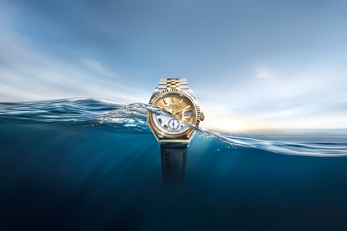 Eine luxuriöse Armbanduhr mit goldenem und silbernem Band taucht aus einem ruhigen, blauen Ozean auf, mit dem Horizont und einem weichen, bewölkten Himmel im Hintergrund. Das Zifferblatt befindet sich teilweise über und teilweise unter dem Wasser.