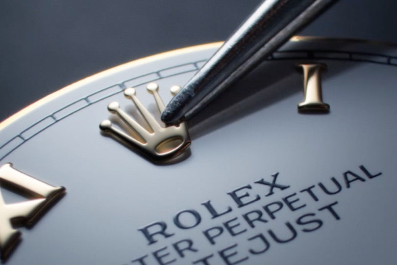 Nahaufnahme eines Rolex-Zifferblatts mit einem Metallwerkzeug, das auf das erhabene goldene Kronenlogo zeigt. Römische Ziffern als Stundenmarkierungen sind teilweise sichtbar, mit den Texten „Rolex“ und „Oyster Perpetual Datejust“ in Schwarz.
