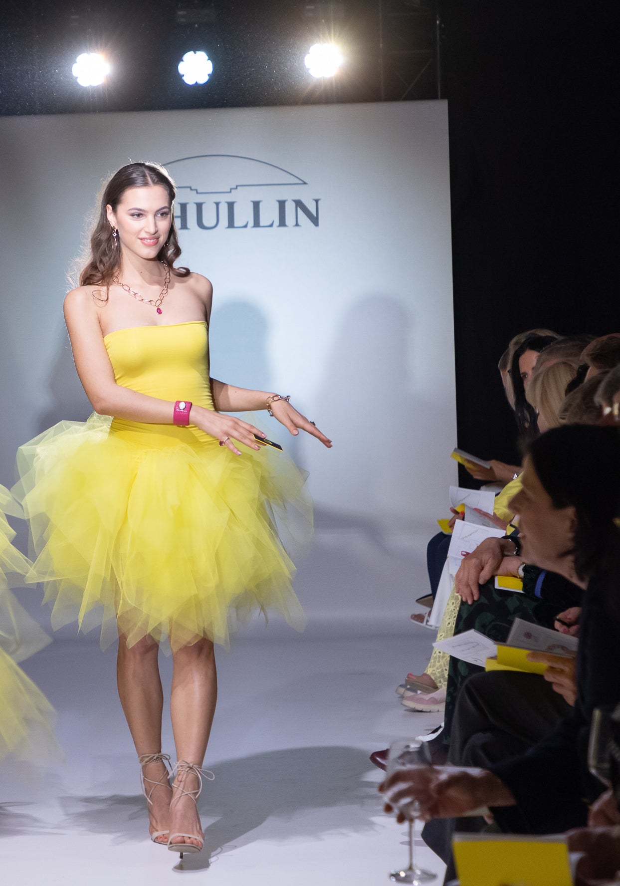 Ein Model in einem gelben trägerlosen Kleid mit Tüllrock läuft über einen Laufsteg. Sie trägt ein Armband und eine Halskette. Das Publikum schaut aufmerksam zu und helle Lichter erhellen die Szene.