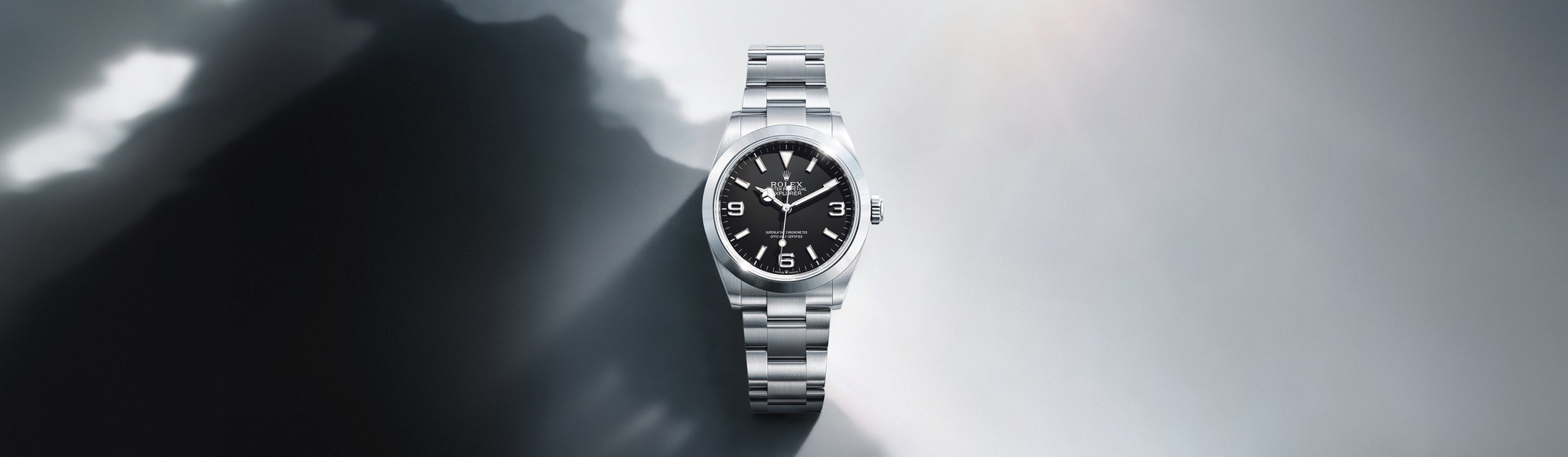 Eine silberne Armbanduhr mit schwarzem Zifferblatt und silbernen Indexen steht im Mittelpunkt eines Hintergrunds mit Farbverlauf, der von dunklen zu hellen Grautönen übergeht, wobei Licht- und Schatteneffekte die Uhr hervorheben.