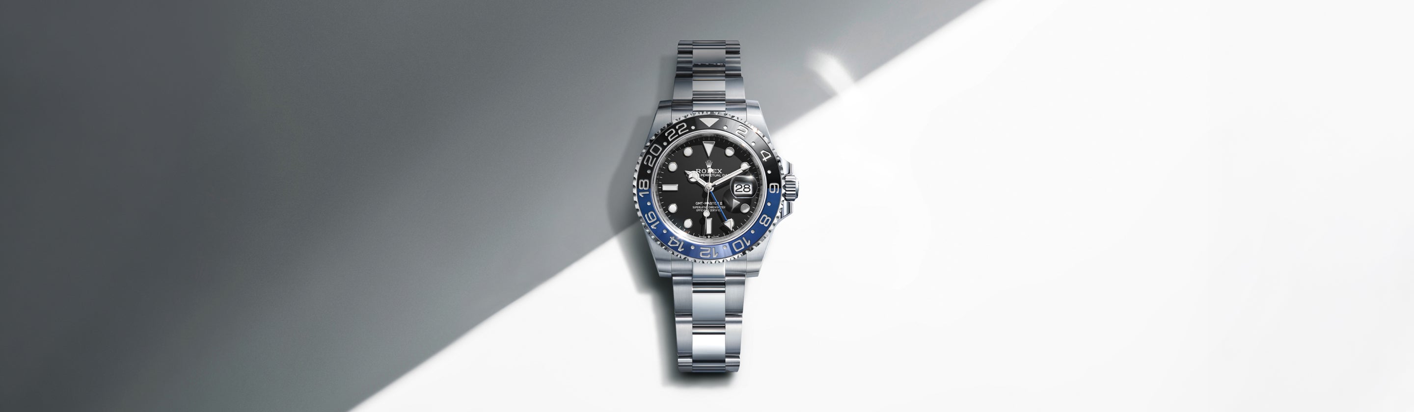 Eine silberne Luxusarmbanduhr mit schwarzer und blauer Lünette, schwarzem Zifferblatt und Metallarmband steht vor einem diagonalen Hintergrund, der zwischen Grau und Weiß aufgeteilt ist.