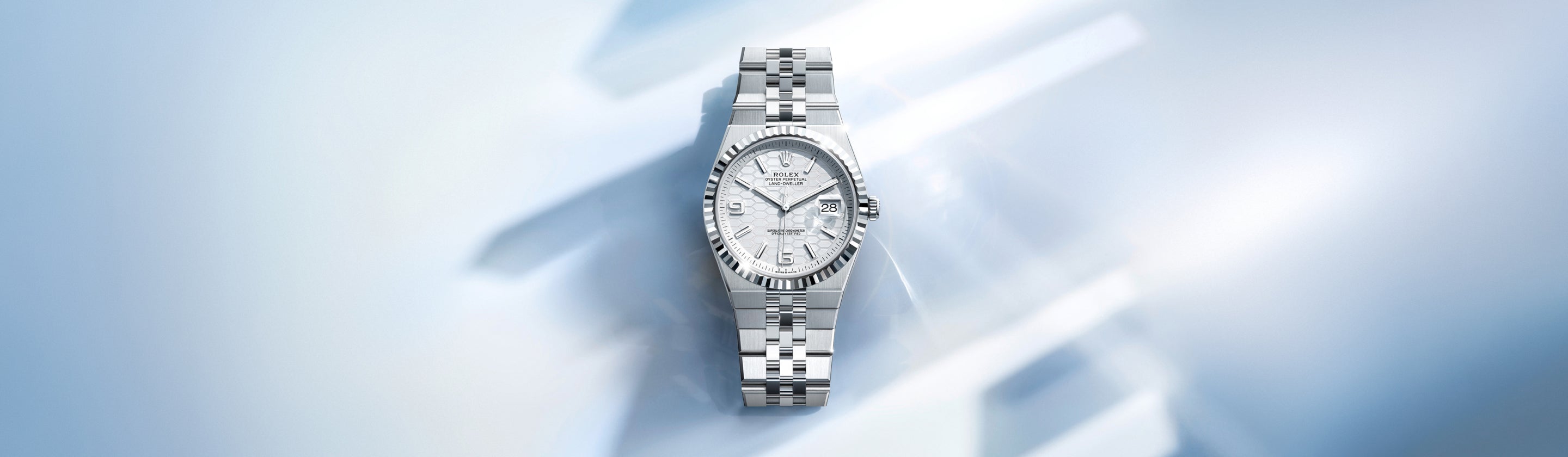 Eine luxuriöse silberne Uhr mit Metallarmband und strukturiertem Zifferblatt wird vor einem weichen, hellblauen, abstrakten Hintergrund mit geometrischen Formen und Reflexionen präsentiert.