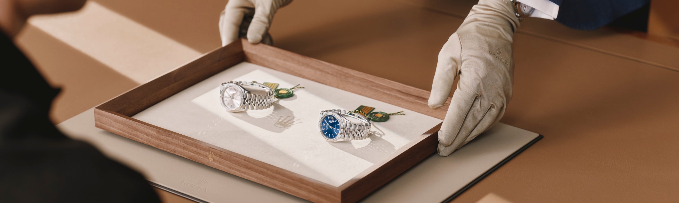 Eine Person mit weißen Handschuhen überreicht einer anderen sitzenden Person ein Holztablett mit zwei Luxusuhren, eine mit einem silbernen und eine mit einem blauen Zifferblatt. Beide Uhren sind mit Metallbändern und Anhängern versehen.