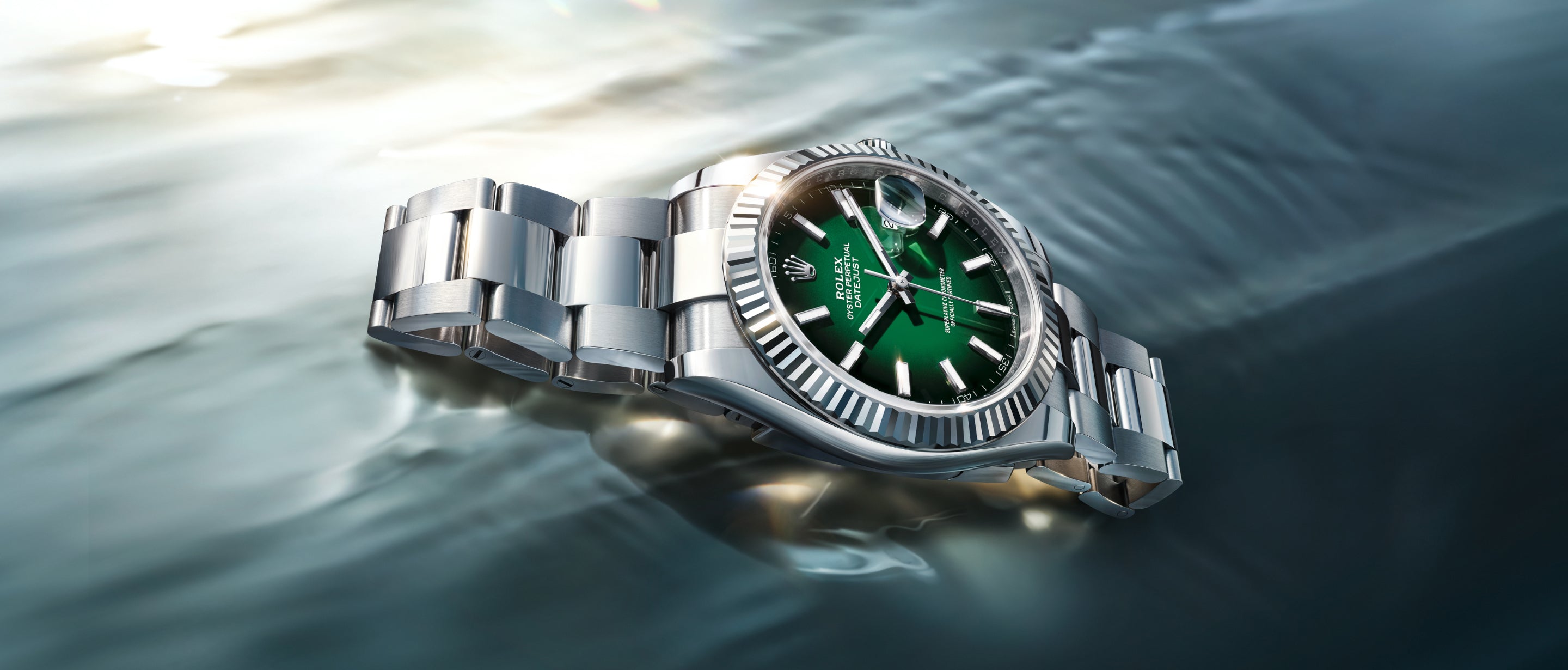 Eine silberne Rolex-Armbanduhr mit grünem Zifferblatt und silbernen Stundenmarkierungen liegt auf einer spiegelnden Wasseroberfläche, die das Licht einfängt und reflektiert.