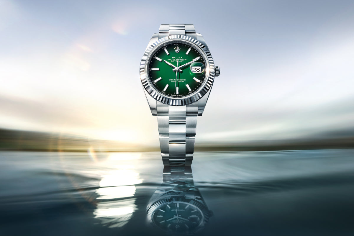 Eine silberne Rolex-Uhr mit grünem Zifferblatt und Datumsanzeige steht aufrecht und spiegelt sich auf einer Wasseroberfläche, vor einem verschwommenen landschaftlichen Hintergrund und Sonnenlicht in der Ferne.