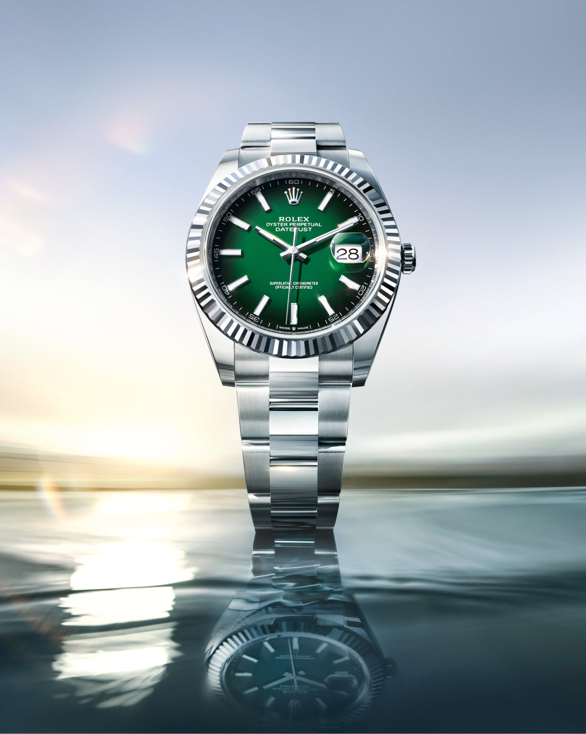 Eine silberne Rolex-Uhr mit grünem Zifferblatt und Datumsanzeige, aufrecht stehend auf einer spiegelnden Oberfläche mit einem weichen, verschwommenen Himmel und einem Wasserhintergrund dargestellt.