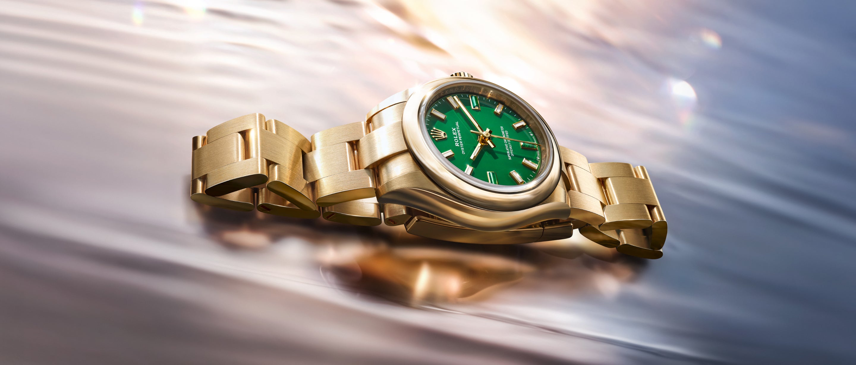 Eine goldene Armbanduhr mit grünem Zifferblatt ist auf einer reflektierenden, gewellten Oberfläche abgebildet. Die Uhr hat ein Metallarmband und wirkt luxuriös, wenn das Licht auf der polierten Oberfläche glitzert.