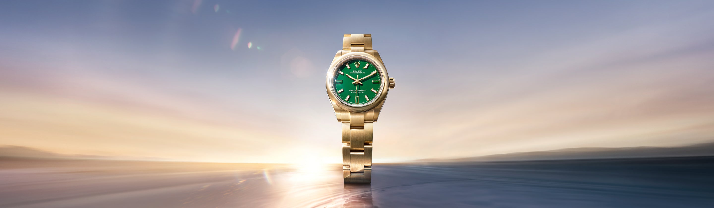 Eine goldene Armbanduhr mit grünem Zifferblatt steht vor einem verschwommenen, verträumten Hintergrund aus sanften Blau-, Gelb- und Rosatönen, der an einen Sonnenauf- oder -untergang erinnert. Das Licht wird von der Uhr und dem Hintergrund reflektiert, wodurch ein Leuchteffekt entsteht.