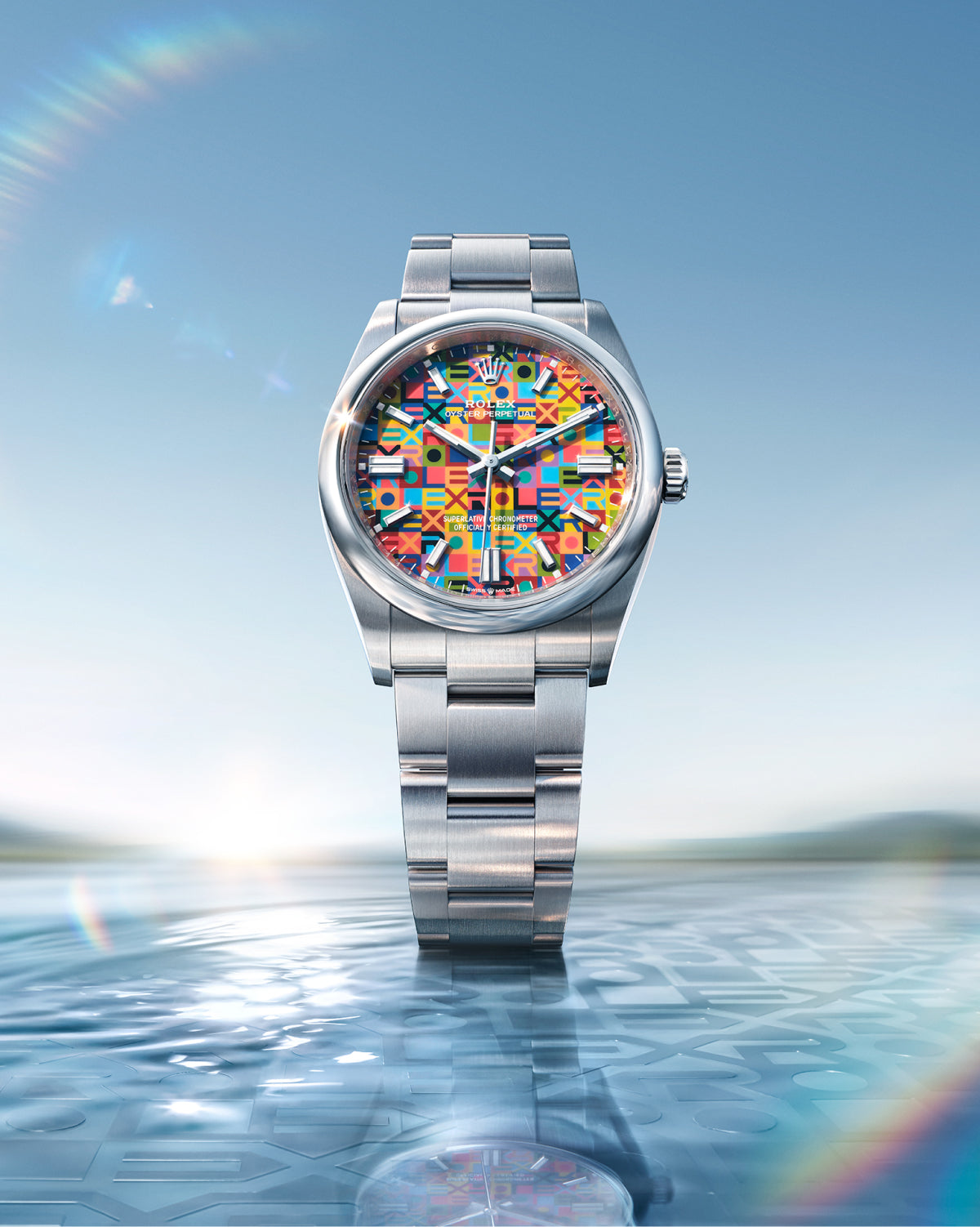 Eine Armbanduhr aus Edelstahl mit einem farbenfrohen, geometrisch gemusterten Zifferblatt steht aufrecht auf einer spiegelnden, wasserähnlichen Oberfläche, mit einem hellen Himmel und einer sanften Landschaft im verschwommenen Hintergrund.