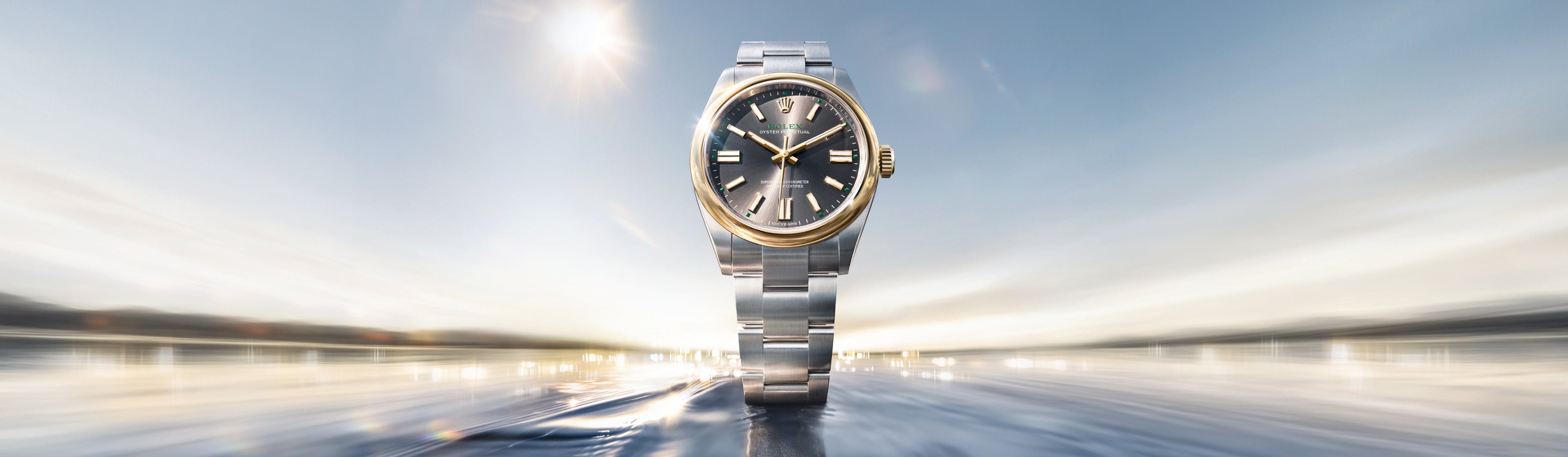 Eine luxuriöse silberne und goldene Armbanduhr mit schwarzem Zifferblatt steht aufrecht auf einer spiegelnden Oberfläche, deren verschwommener, sonnenbeschienener Hintergrund Bewegung und Eleganz suggeriert.
