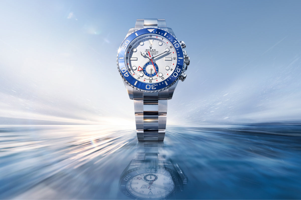 Eine luxuriöse silberne Rolex-Uhr mit blau-weißem Zifferblatt und Lünette wird aufrecht präsentiert und spiegelt sich auf einer glänzenden blauen Oberfläche mit einem weichen, verschwommenen Hintergrund, der an Geschwindigkeit und Wasser erinnert.