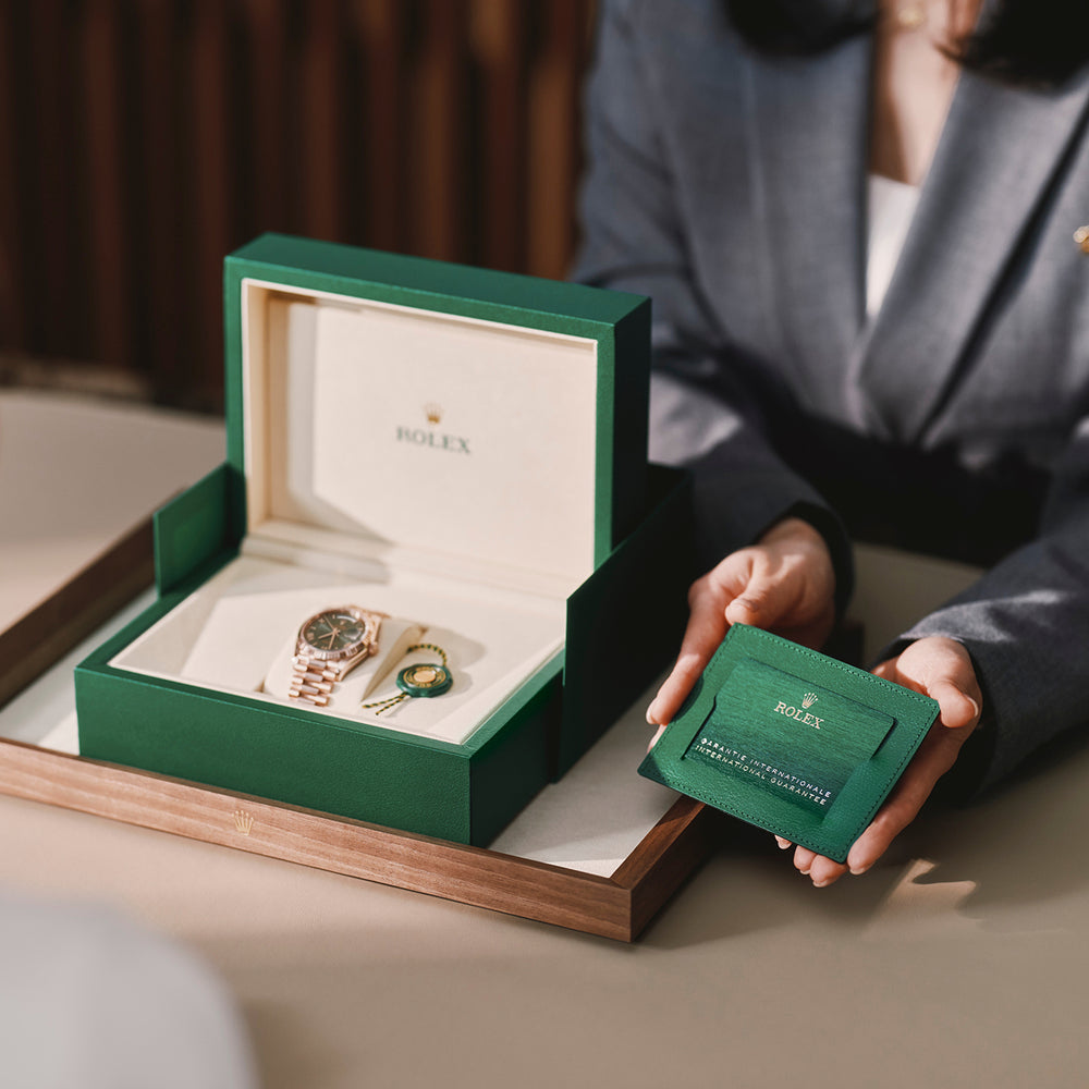 Eine Person im Anzug präsentiert eine grüne Rolex-Schachtel mit einer goldenen Uhr, einem Anhänger und einem grünen Kartenhalter mit einer Garantiekarte auf einem beigen Tisch.
