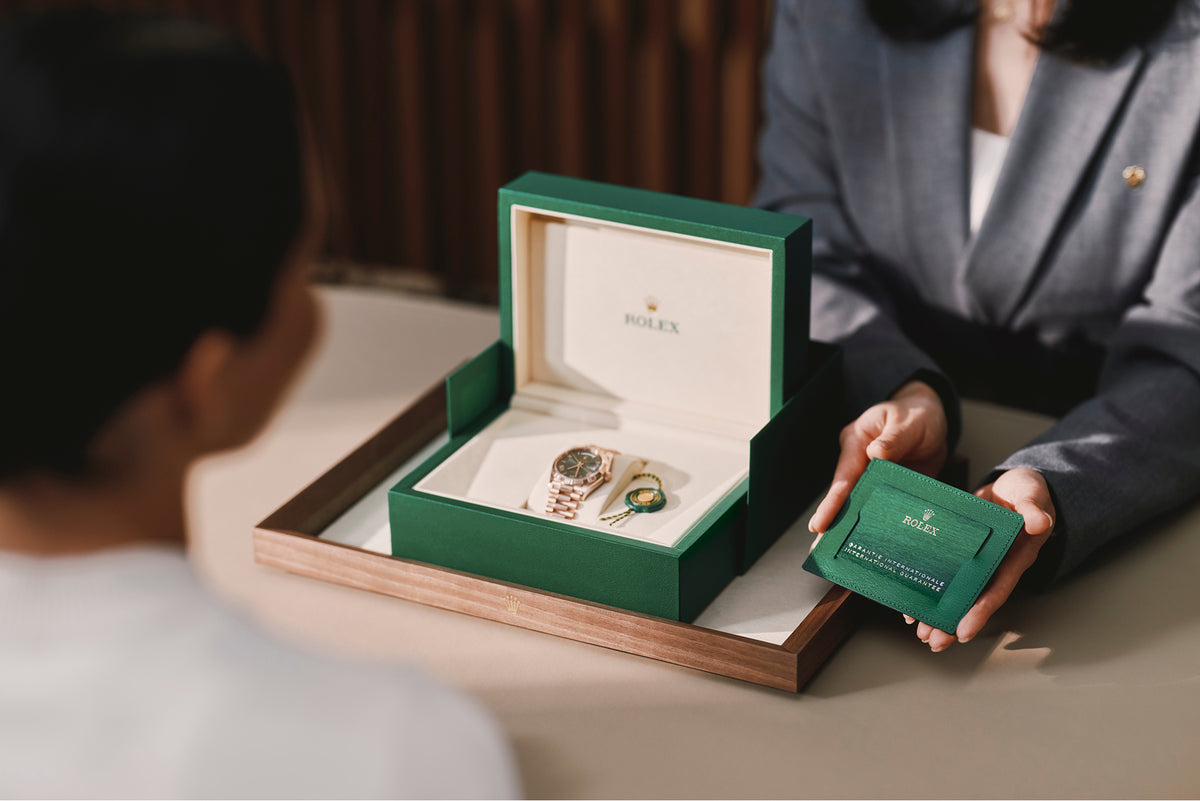 Eine Person überreicht einer anderen Person an einem Tisch eine Rolex-Uhr in einer grünen Schachtel und eine grüne Brieftasche mit Dokumenten. Beide sitzen, und nur ihre Oberkörper und Hände sind sichtbar.