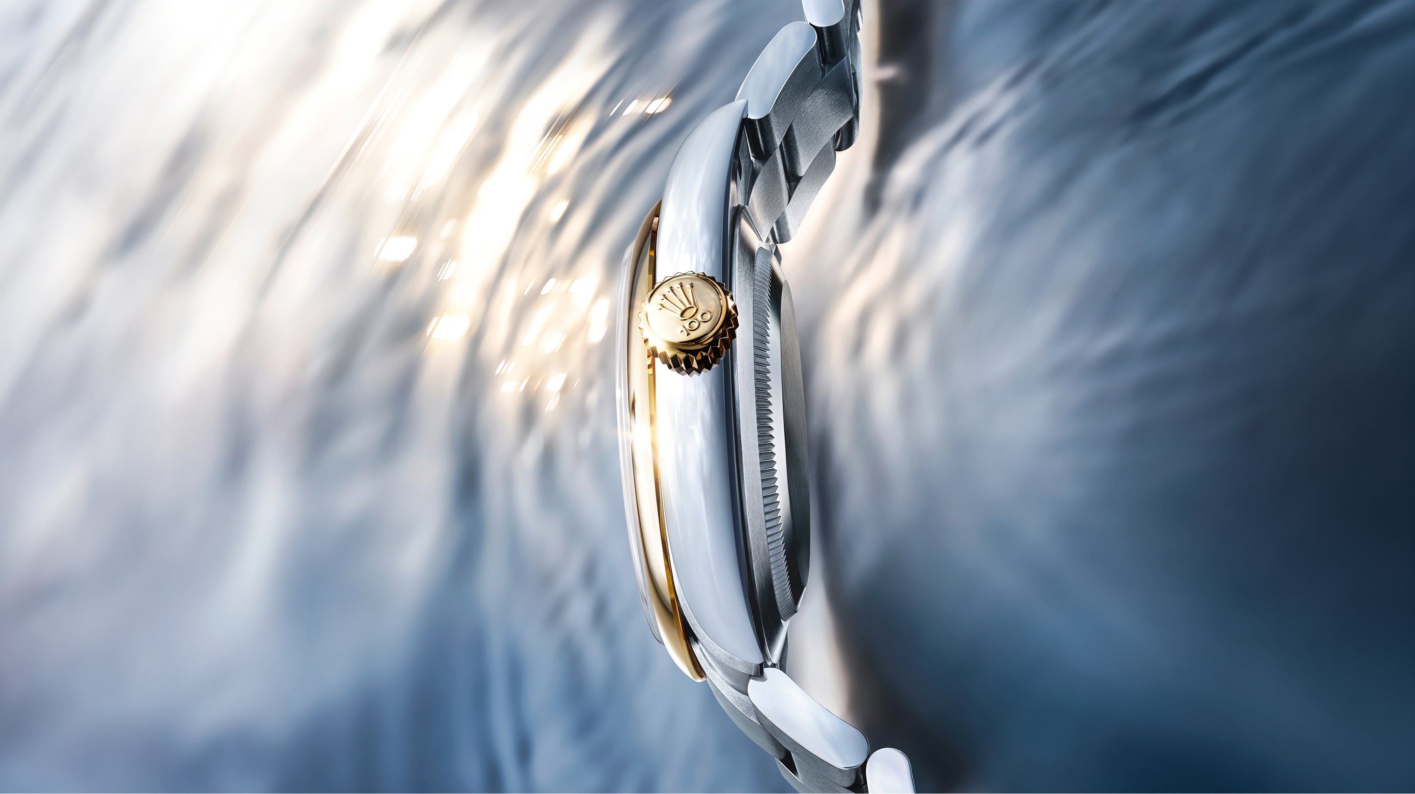 Nahaufnahme einer Luxusarmbanduhr mit goldener Krone, silbernem Gehäuse und Metallarmband vor einem sanft verschwommenen, reflektierenden Hintergrund mit schimmerndem Licht.