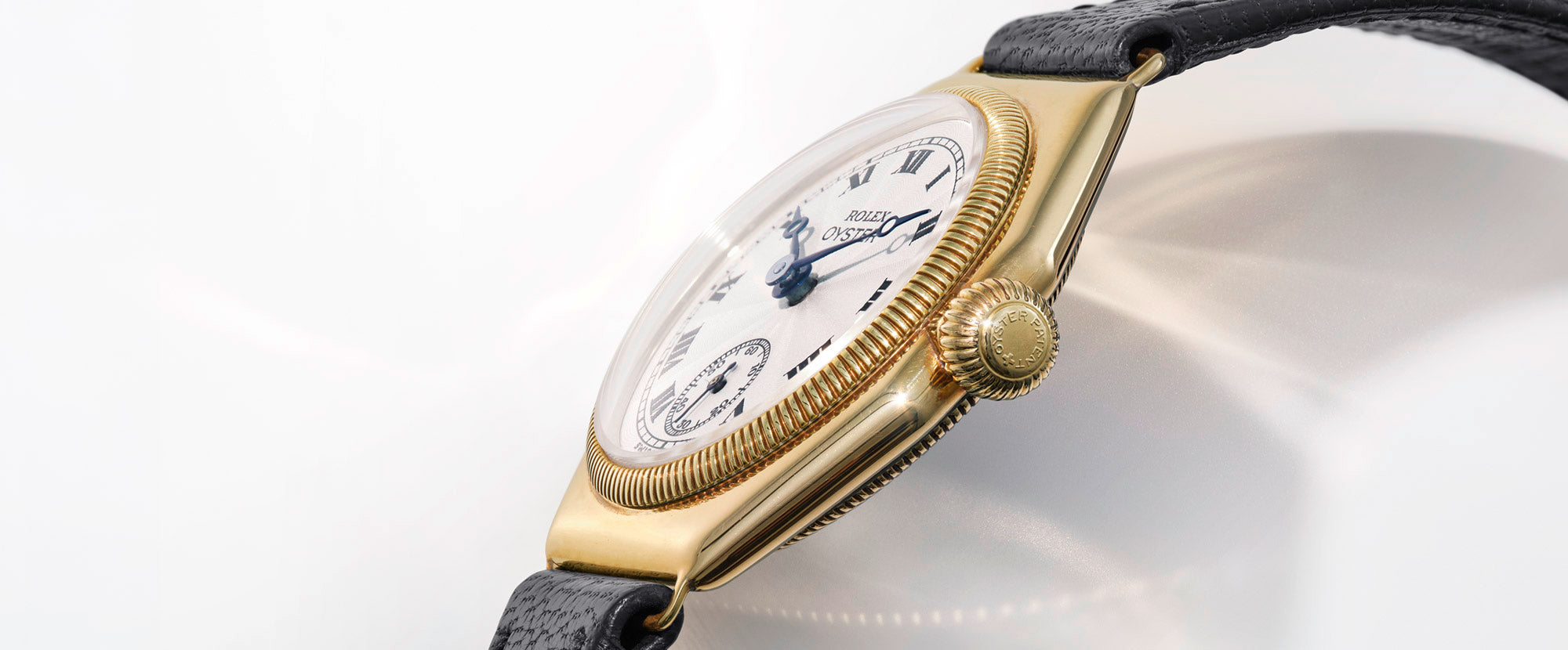 Seitliche Nahaufnahme einer goldenen Armbanduhr mit weißem Zifferblatt, schwarzem Lederarmband und römischen Ziffern, die mit komplizierten Details und einer geriffelten Krone versehen ist.