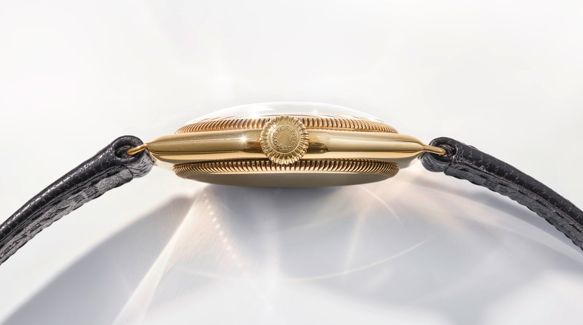 Seitenansicht einer goldenen Armbanduhr mit strukturierter Krone und schwarzem Lederarmband, die auf einer hellen Oberfläche mit weichen Reflexen und Schatten liegt.