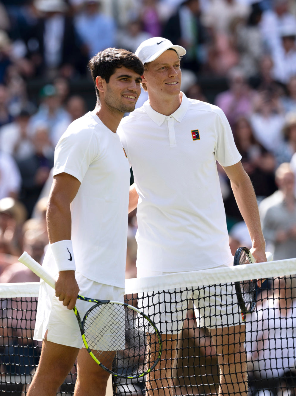 Zwei männliche Tennisspieler in weißen Outfits stehen nebeneinander am Netz und lächeln für ein Foto vor einem Spiel. Beide halten Schläger in der Hand, und im Hintergrund ist eine Menge von Zuschauern zu sehen.