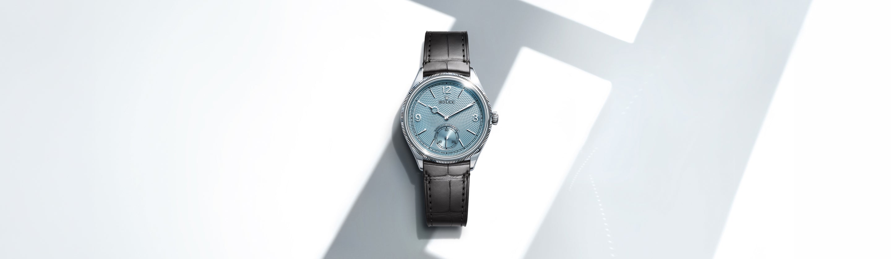 Eine luxuriöse Armbanduhr mit hellblauem, strukturiertem Zifferblatt und schwarzem Lederarmband wird auf einer weißen Fläche mit geometrischen Schatten dargestellt.