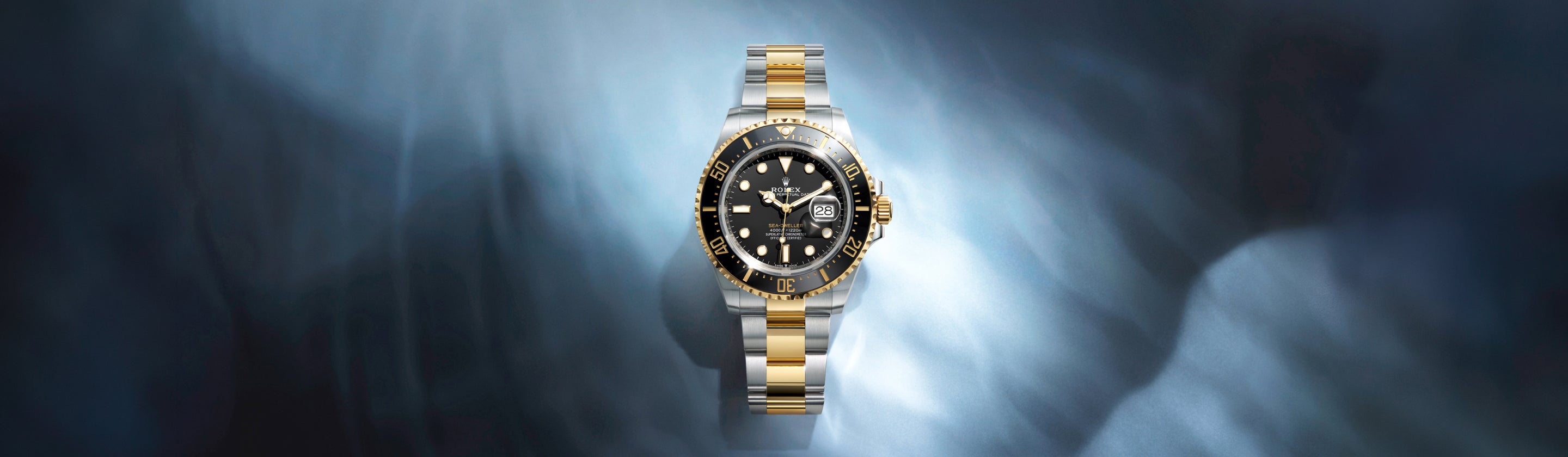 Eine luxuriöse, zweifarbige Armbanduhr mit schwarzem Zifferblatt und goldenen Akzenten ist auf einem metallischen, verschwommenen blauen Hintergrund zentriert, hinter dem Licht strahlt.