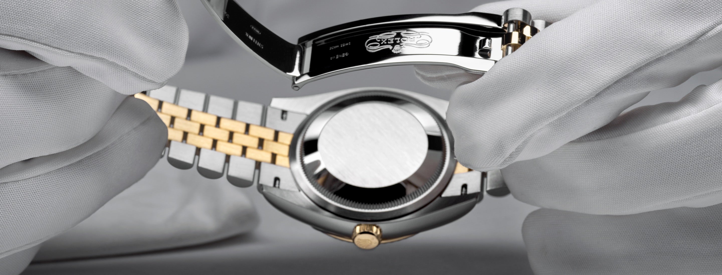 Eine Nahaufnahme einer Person mit weißen Handschuhen, die eine Luxusarmbanduhr mit einem goldenen und silbernen Armband hält. Die Rückseite der Uhr ist offen und gibt einen Teil ihres Mechanismus frei. Das Innere der Schließe ist sichtbar.