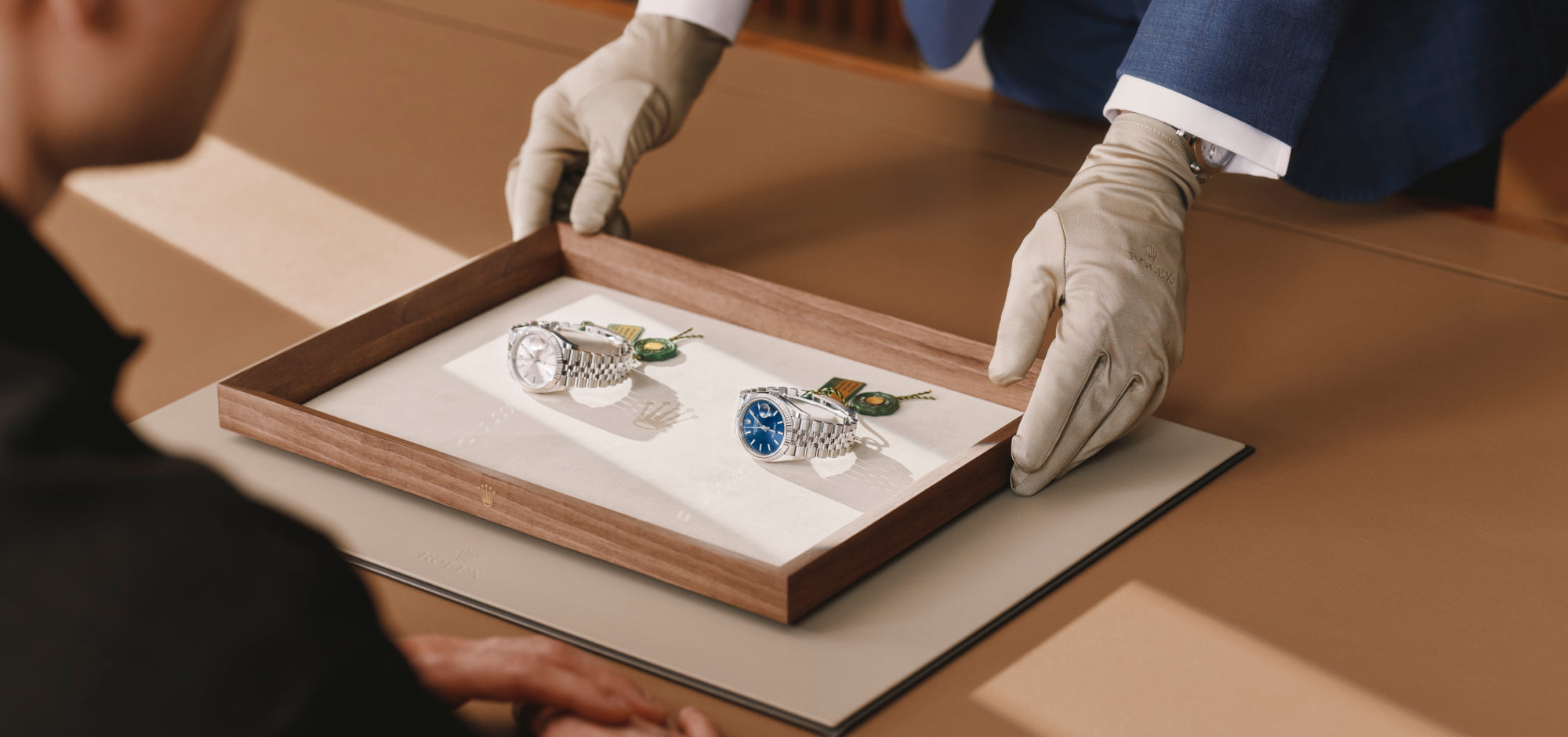 Eine Person mit Handschuhen überreicht einer anderen Person an einem gut beleuchteten, eleganten Tisch ein Holztablett mit zwei Luxusarmbanduhren auf einer weichen Matte.