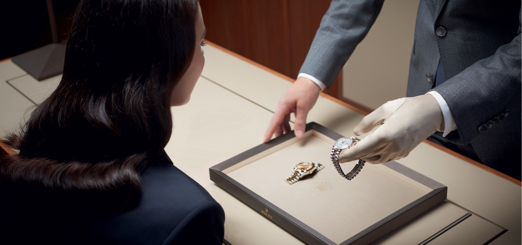 Eine Person mit Handschuhen überreicht einem sitzenden Kunden in einem Luxusgeschäft eine Luxusuhr. Eine zweite Uhr liegt in einer Vitrine auf dem Tisch. Die Atmosphäre ist formell und elegant.