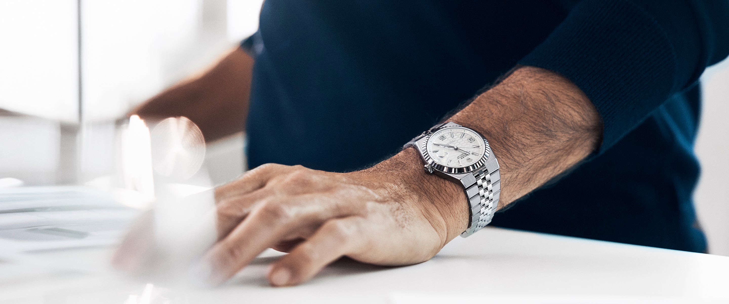 Nahaufnahme von Unterarm und Hand einer Person, die auf einem weißen Tisch liegt und eine silberne Armbanduhr mit Metallband und weißem Zifferblatt trägt. Die Person ist mit einem dunklen, langärmeligen Hemd bekleidet.