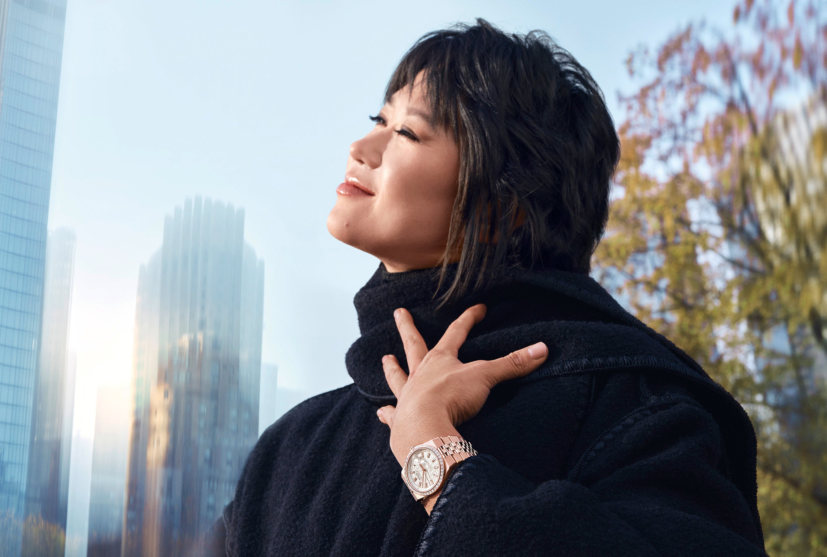 Eine Frau in einem schwarzen Mantel lächelt mit geschlossenen Augen, berührt ihren Hals und zeigt eine elegante Armbanduhr. Hohe Gebäude und Bäume sind im unscharfen Hintergrund unter einem klaren Himmel zu sehen.
