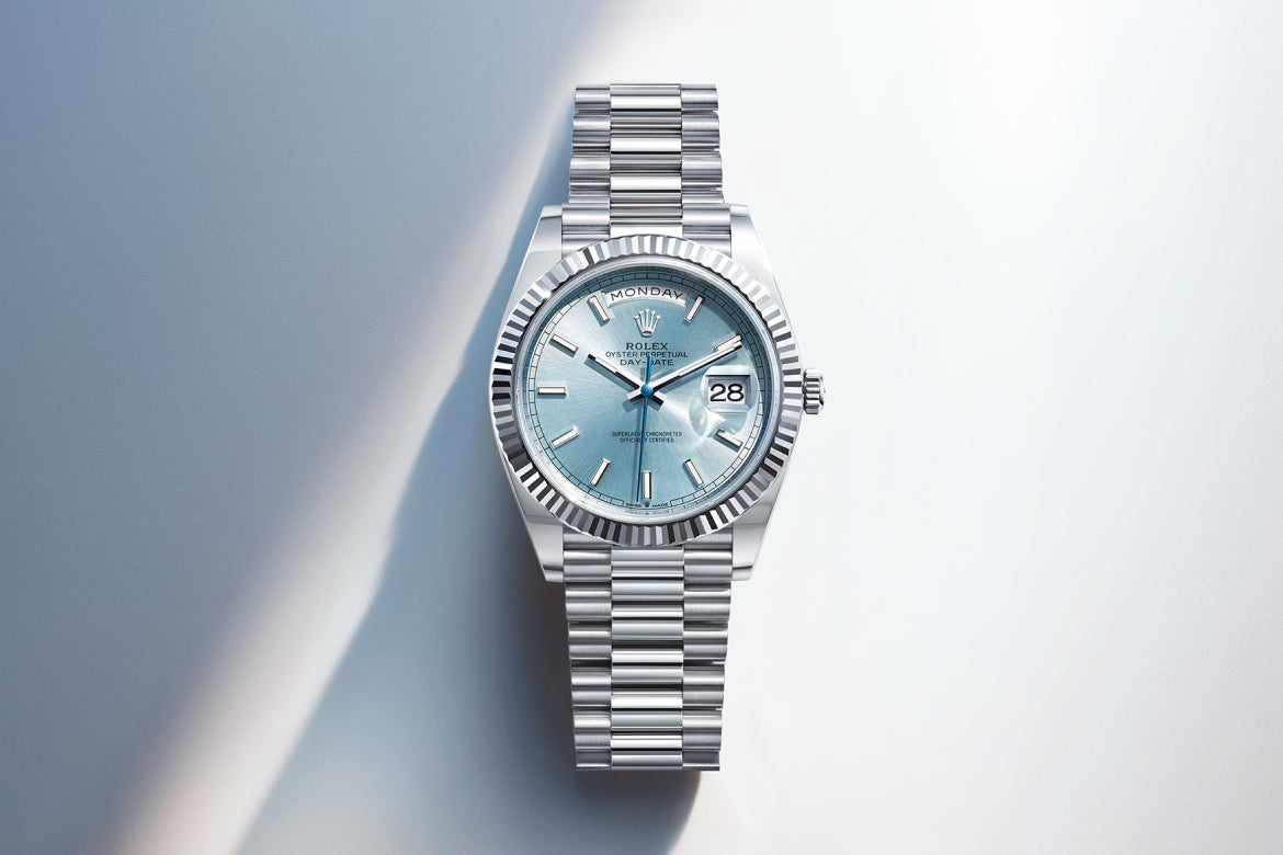 Eine silberne Rolex-Armbanduhr mit hellblauem Zifferblatt, geriffelter Lünette und Metallgliederarmband, die den Tag und das Datum anzeigt, fotografiert auf einem hellen Hintergrund mit Farbverlauf.