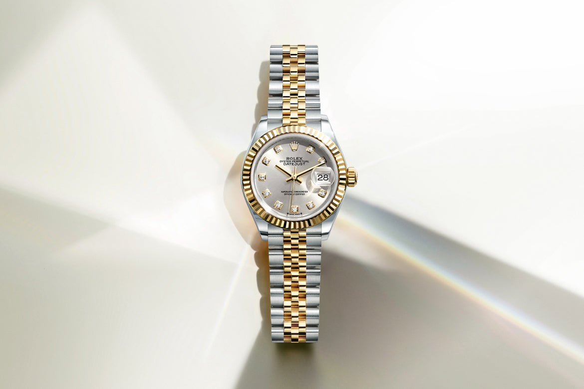 Eine silber- und goldfarbene Luxus-Armbanduhr mit geriffelter Lünette, silbernem Zifferblatt, diamantbesetzten Indexen und zweifarbigem Metallarmband auf hellem Hintergrund mit sanften Reflexen.