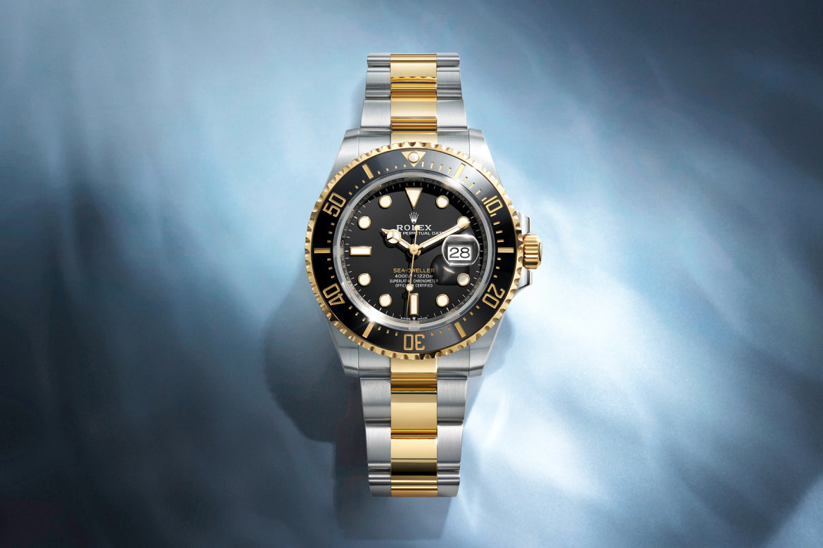 Eine zweifarbige Rolex-Uhr mit schwarzem Zifferblatt und schwarzer Lünette, goldenem und silbernem Armband und einer Datumsanzeige, dargestellt vor einem Hintergrund mit blauem und weißem Farbverlauf.