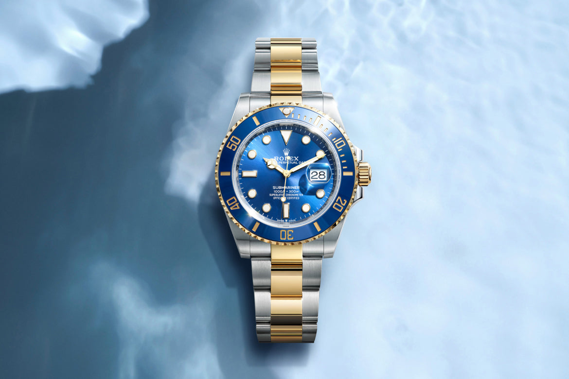 Eine zweifarbige Rolex-Uhr in Gold und Silber mit blauem Zifferblatt und Lünette wird auf einem hellblauen, wasserähnlichen Hintergrund angezeigt. Die Uhr zeigt das Datum und verfügt über leuchtende Stundenmarkierungen.