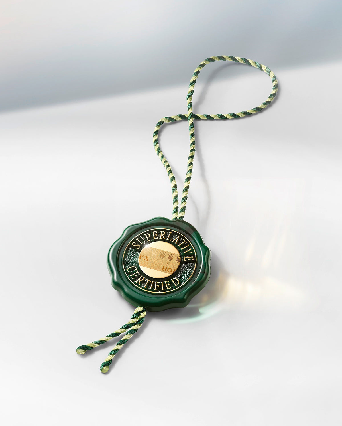 Ein grün-goldenes Rolex "Superlative Certified"-Anhängerschild mit gedrehter grün-weißer Schnur liegt auf einer glatten, reflektierenden weißen Oberfläche.