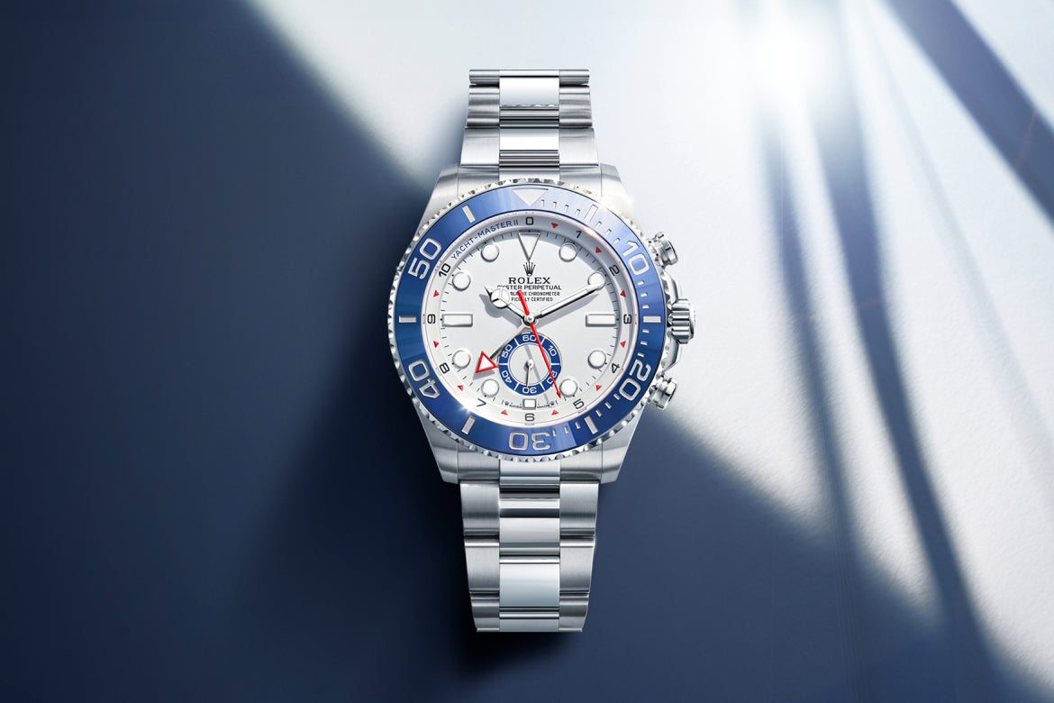 Eine luxuriöse Rolex Yacht-Master II Uhr mit silbernem Metallband und weißem Zifferblatt, akzentuiert durch eine blaue Keramiklünette, dargestellt auf einem blauen und weißen Hintergrund mit weichen, schrägen Schatten.