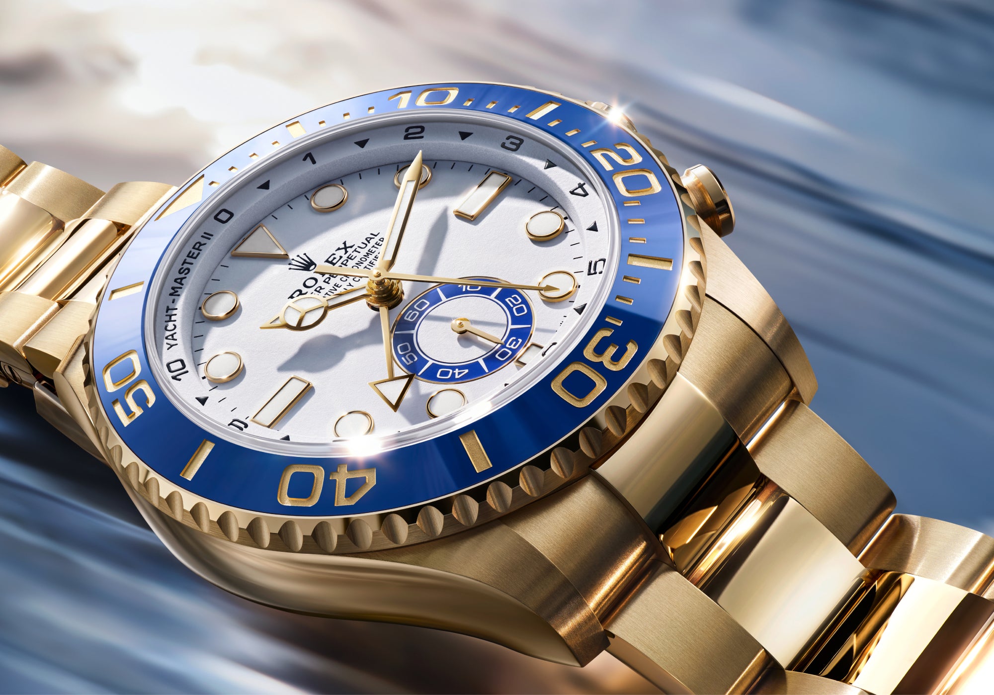 Eine Nahaufnahme einer goldenen Rolex Yacht-Master II Uhr mit weißem Zifferblatt, blauer Lünette und goldenem Armband vor einem unscharfen blauen und weißen Hintergrund.