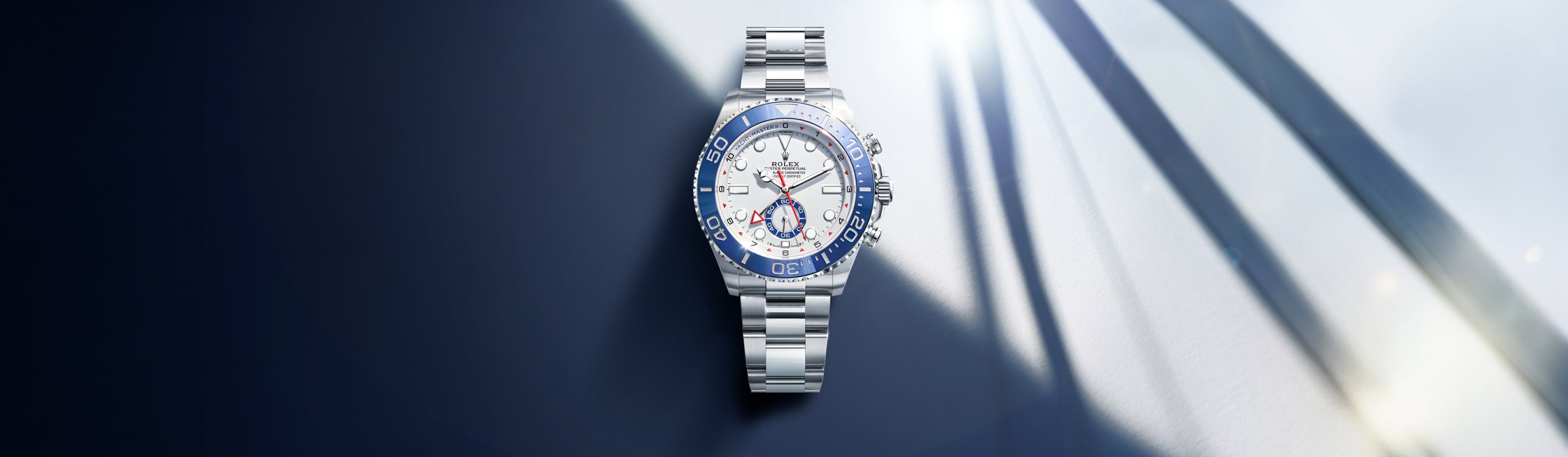 Eine luxuriöse silberne Uhr mit blau-weißem Zifferblatt und Lünette wird auf einer spiegelnden Oberfläche mit dramatischen Schatten und von rechts einfallendem Licht dargestellt.