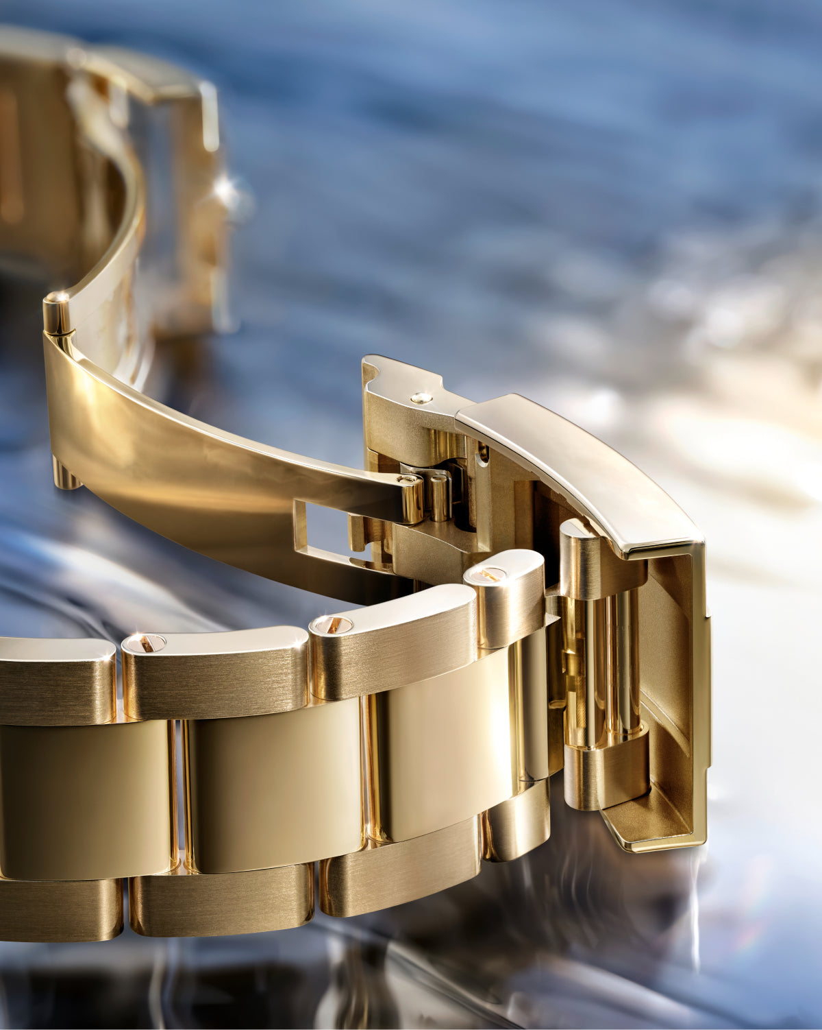 Nahaufnahme eines goldenen Metallarmbands mit teilweise geöffnetem Verschluss, dargestellt auf einer spiegelnden Oberfläche mit einem unscharfen blauen und silbernen Hintergrund.