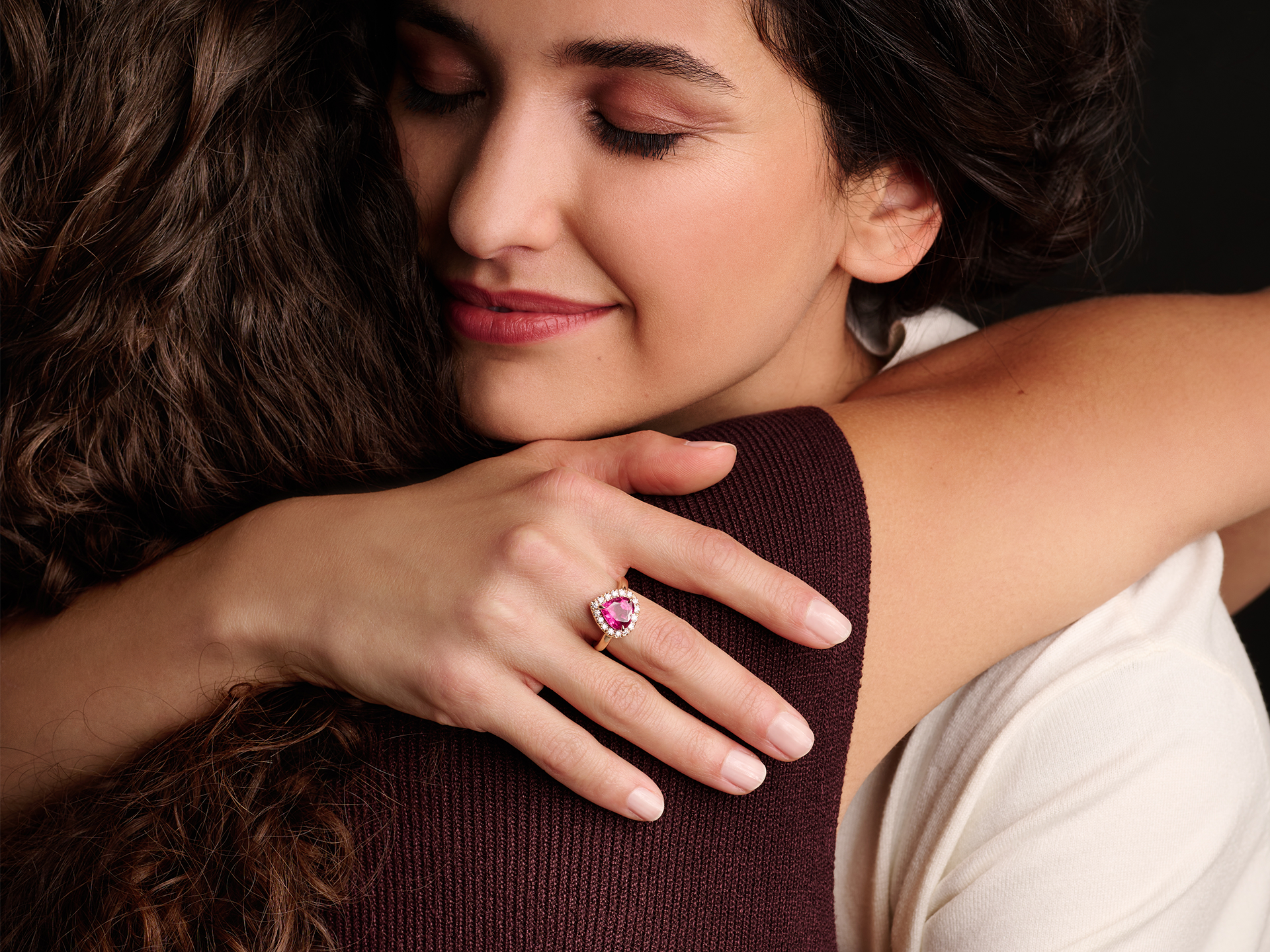 Eine Frau mit dunklem gewelltem Haar lächelt sanft, die Augen geschlossen, während sie jemanden umarmt. An ihrer Hand ruht der Schullin HERZ RING CARMOISIERT, ein großer rosa Edelsteinring.