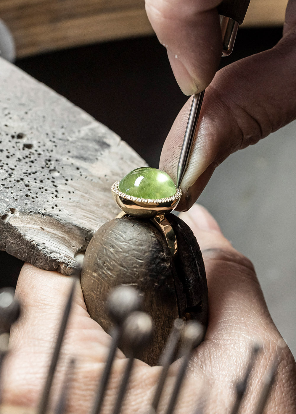 Die Hand eines Juweliers setzt mithilfe eines kleinen Werkzeugs einen runden grünen Edelstein in einen Goldring ein. Der Ring wird sicher in einer Holzklammer auf einer Werkbank gehalten, umgeben von verschiedenen Werkzeugen.